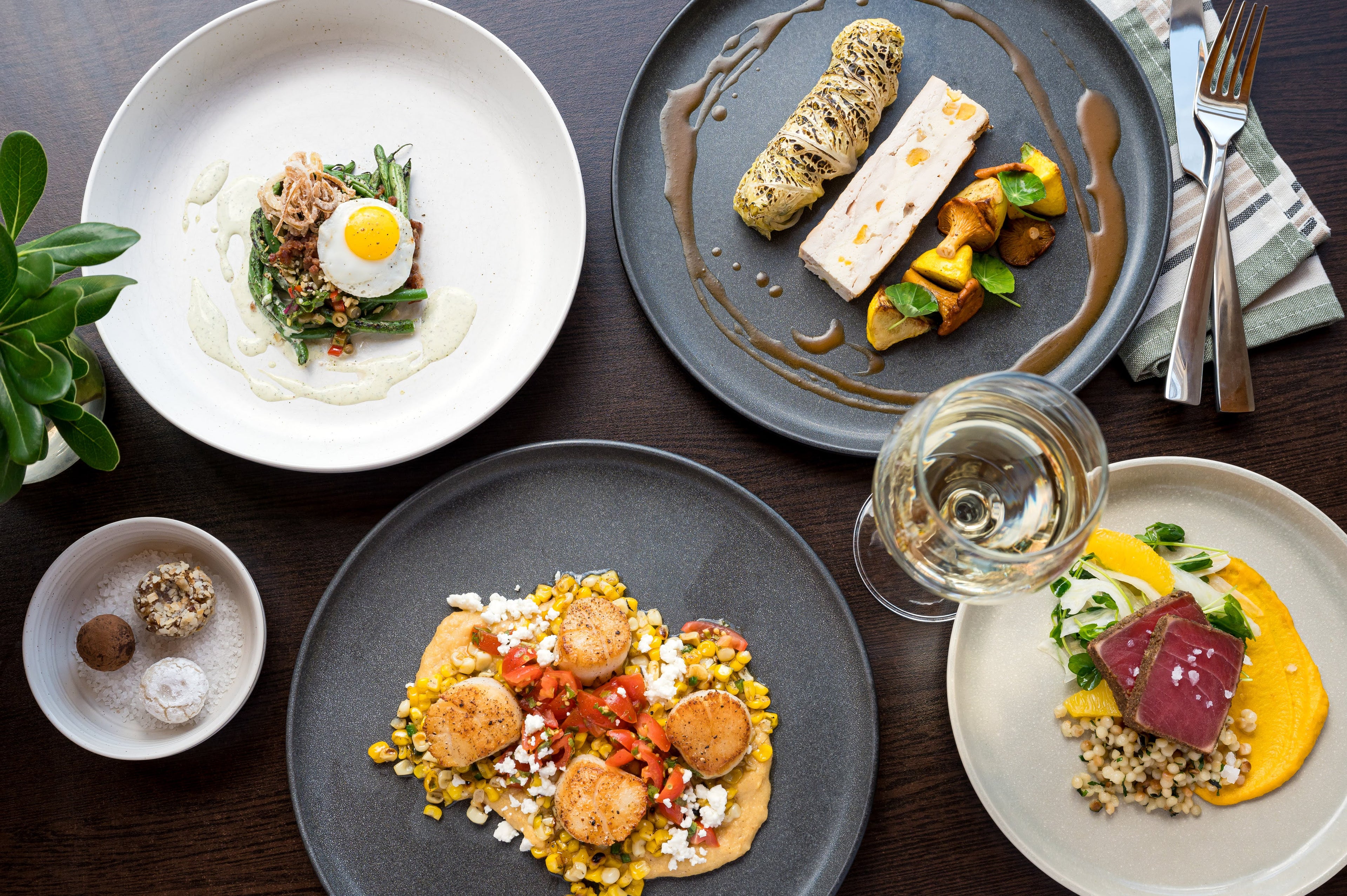 Assorted dishes of gourmet food on a dark wooden table with a glass of white wine.
