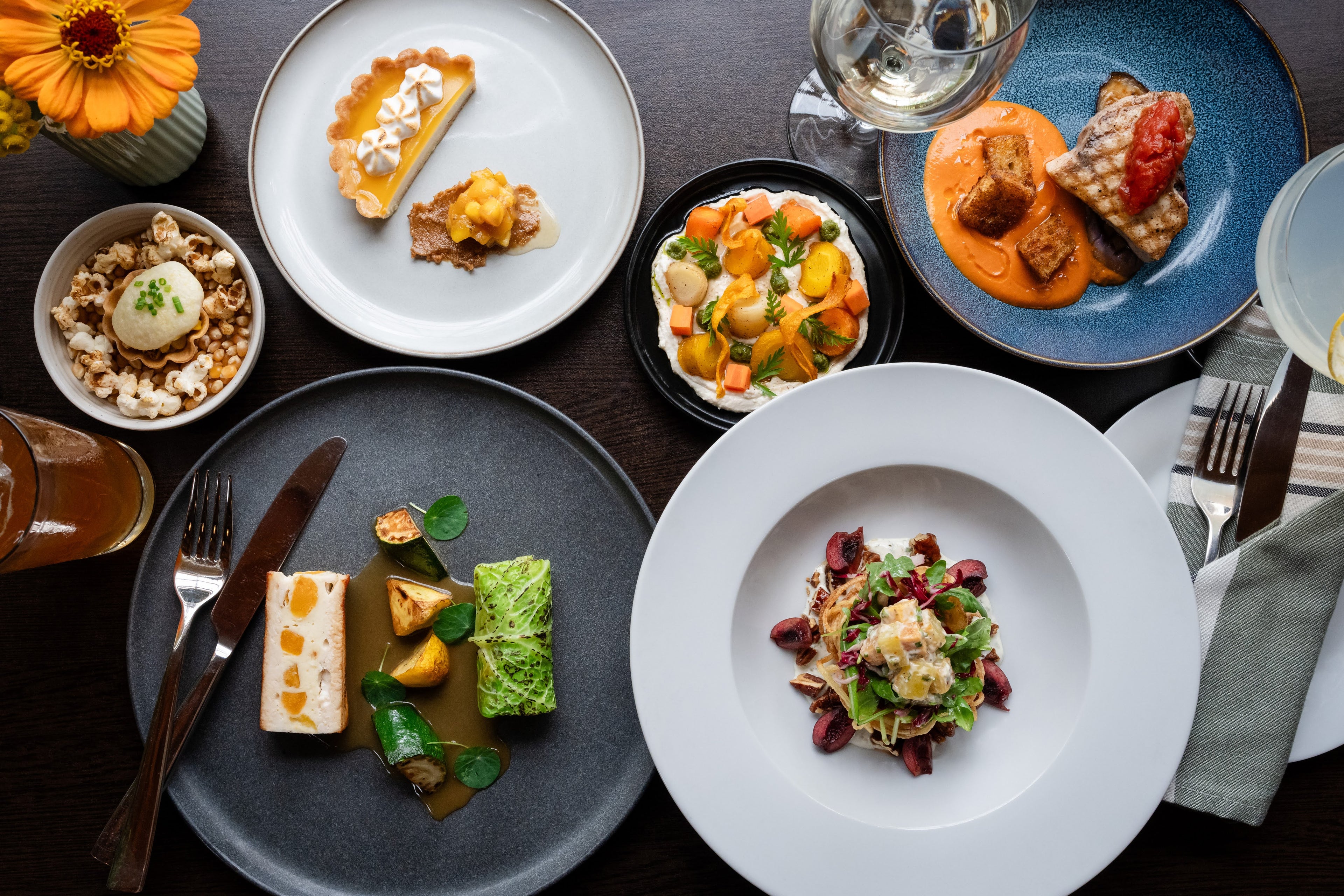 Assorted plates of gourmet food on a dark surface with wine glasses.