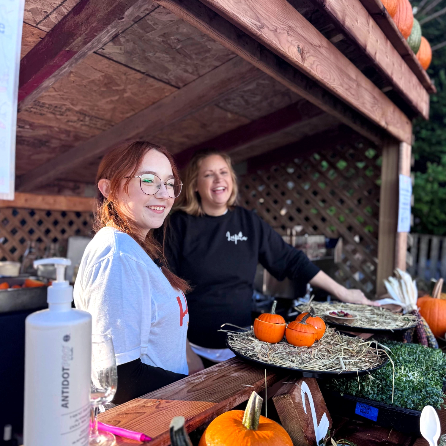 Two women standing behind a counter with pumpkins and other items at an outdoor wooden booth.