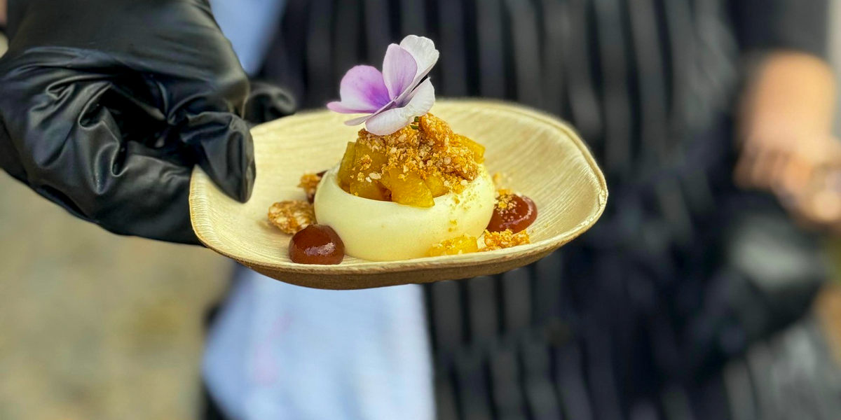 A small plated dessert topped with an edible purple flower, presented in a gloved hand.