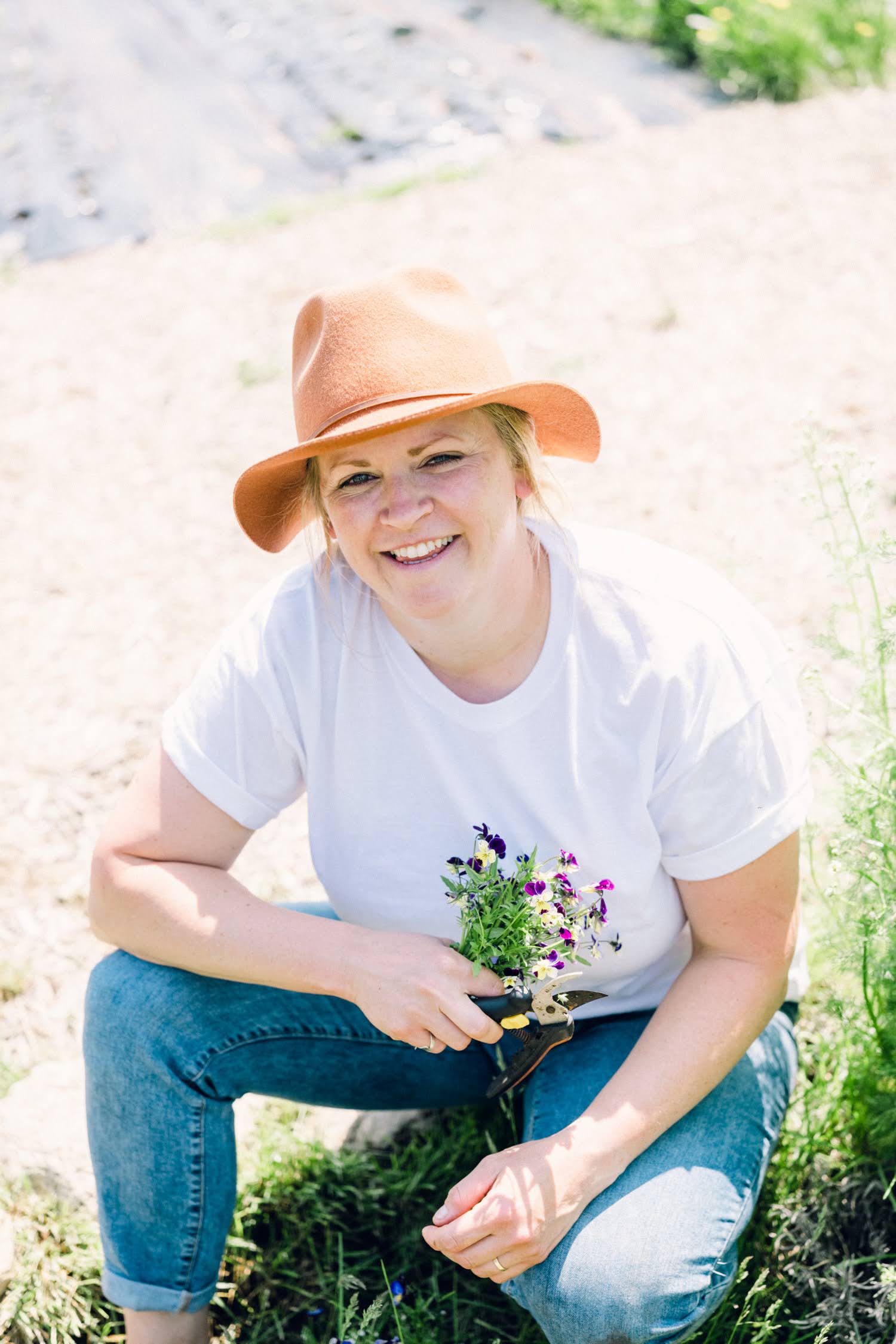 Chef Kim wearing a white t-shirt, blue jeans, and an orange hat, holding flowers outdoors.